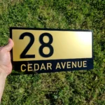 Hand holding a black and gold personalised acrylic house number sign displaying 28 Cedar Avenue outdoors.